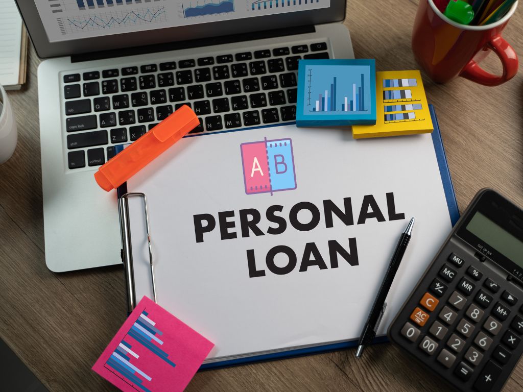 A desk setup with a clipboard labeled "Personal Loan," a laptop, calculator, charts, and office supplies, symbolizing loan analysis and financial planning.